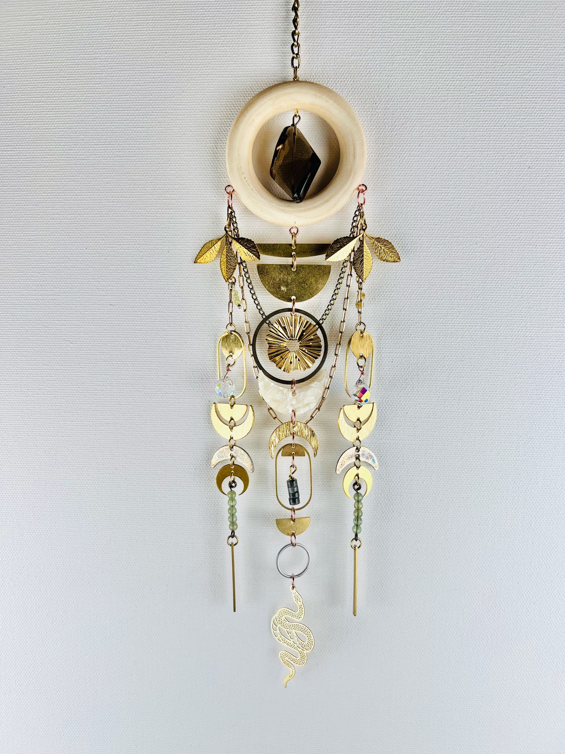 Decorative hanging crystal wall art with gold leaves, half cirlces, rings, ovals, crescent moons, a snake, chain and smoky quartz, green garnet and eagle eye beads and stones on a light gray background