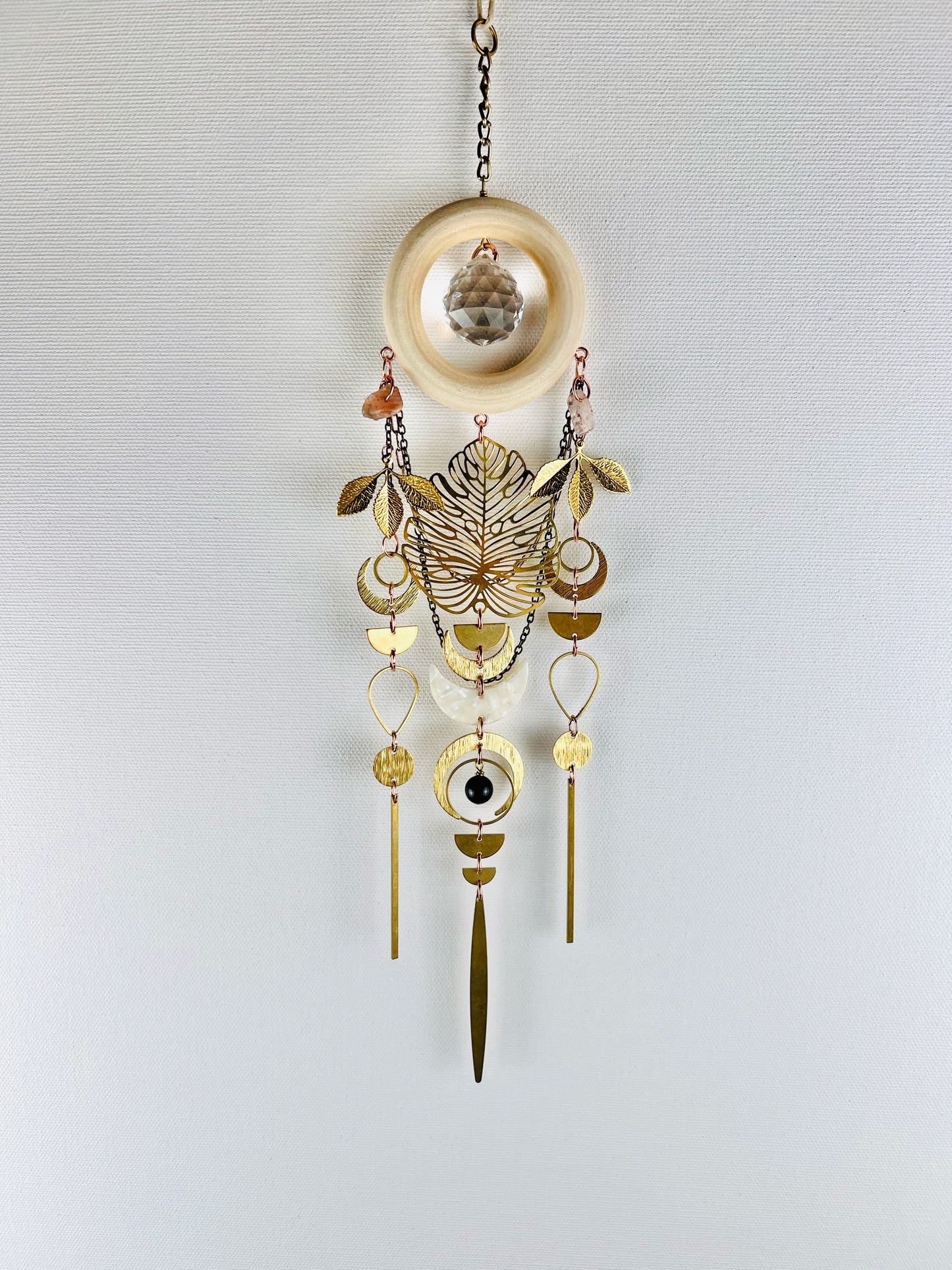 Decorative hanging suncatcher with prism, gold leaves, crescent moons, sunstone, drops, circles, chain and wood ring on a light gray background