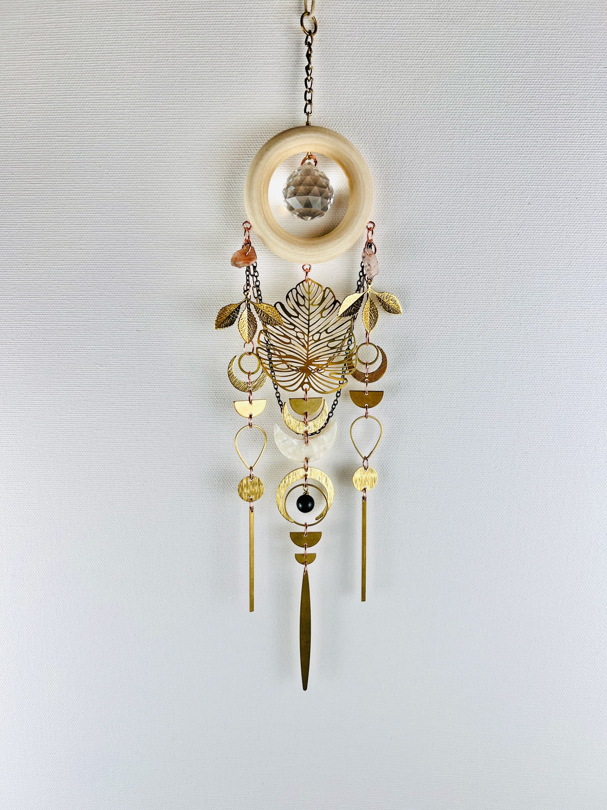 Decorative hanging suncatcher with prism, gold leaves, crescent moons, sunstone, drops, circles, chain and wood ring on a light gray background