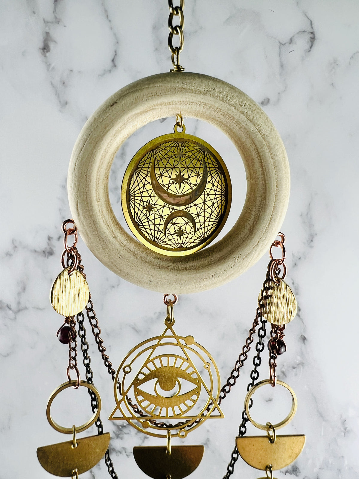 Hanging crystal wall art, including a wood ring on gold medal chain with metal components, including the evil eye, gold rings, half circles, and Drop shapes with metal chain in the background in front of a marble wall.