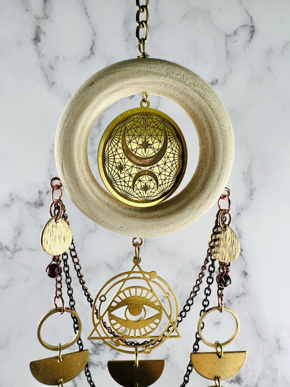 Hanging crystal wall art, including a wood ring on gold medal chain with metal components, including the evil eye, gold rings, half circles, and Drop shapes with metal chain in the background in front of a marble wall.
