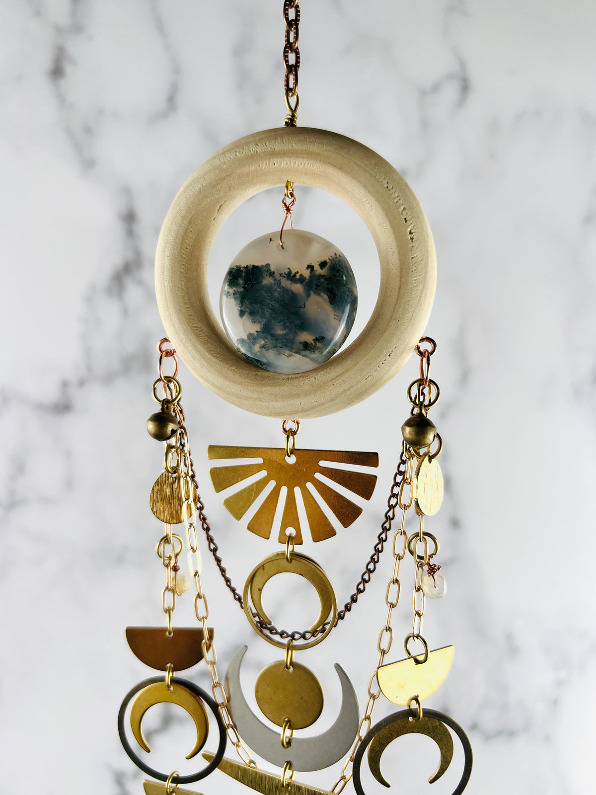 a light brown wood ring hanging from a gold colored chain with a center stone of green and off white moss agate. Below the ring hangs swagging gold and bronze colored chain and metal shapes including: an upside down sunburst, brass bells, wire wrapped topaz beads, a silver crescent moon, gold crescent moons, half circles and rings, hanging in a connected vertical pattern in front of a marble wall. 