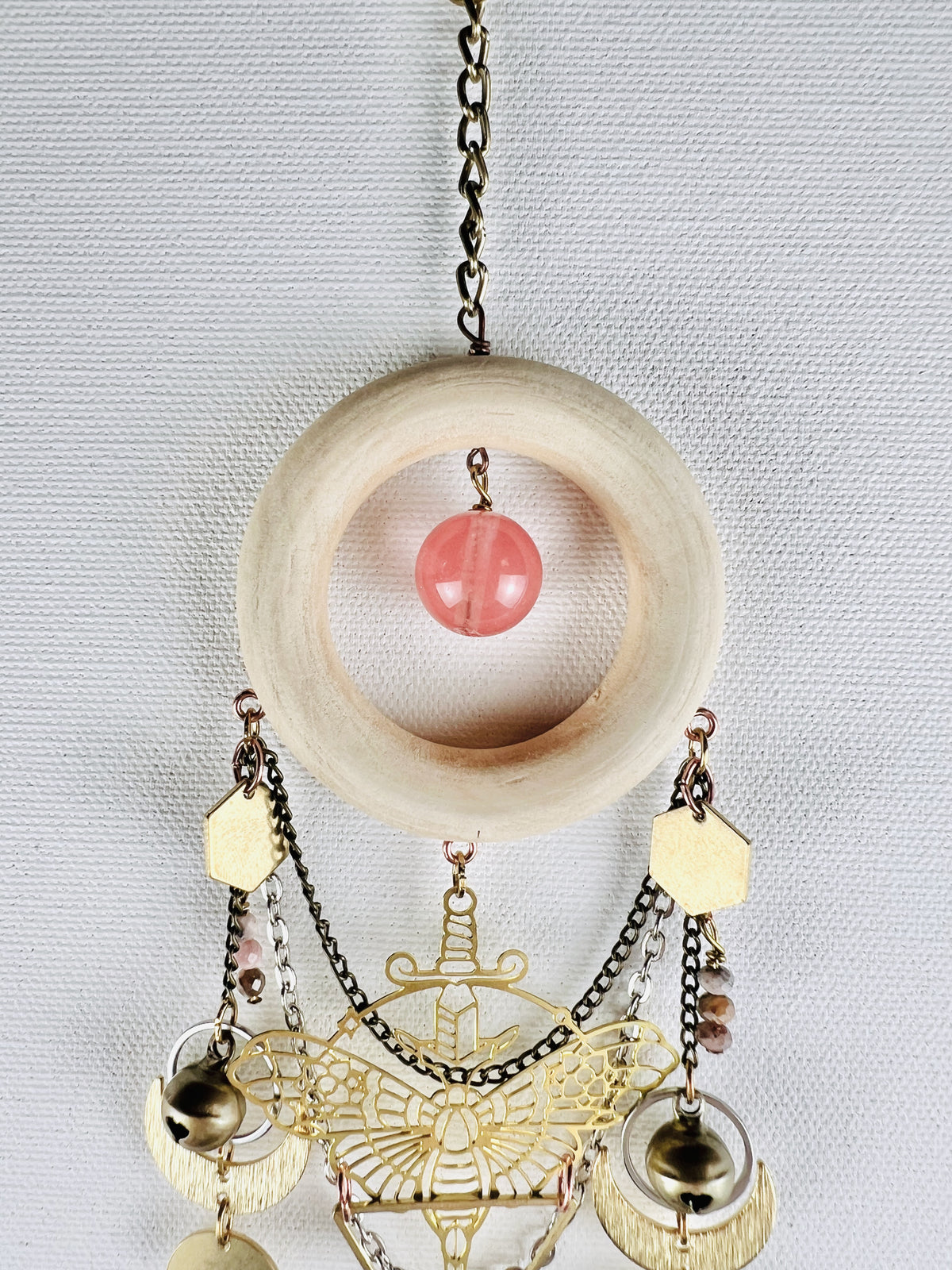 Bronze's colored chain attached to a cream colored wood ring with a cherry quartz round hanging in the center. Attached to that, hanging vertically below, are bronze and silver chains, a gold colored metal moth charm with a sword, rhodochrosite beads, bells, silver rings, crescent moons, and the top of an upside down triangle. All hang in front of a light gray canvas wall.