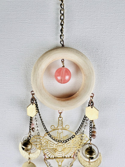 Bronze's colored chain attached to a cream colored wood ring with a cherry quartz round hanging in the center. Attached to that, hanging vertically below, are bronze and silver chains, a gold colored metal moth charm with a sword, rhodochrosite beads, bells, silver rings, crescent moons, and the top of an upside down triangle. All hang in front of a light gray canvas wall.