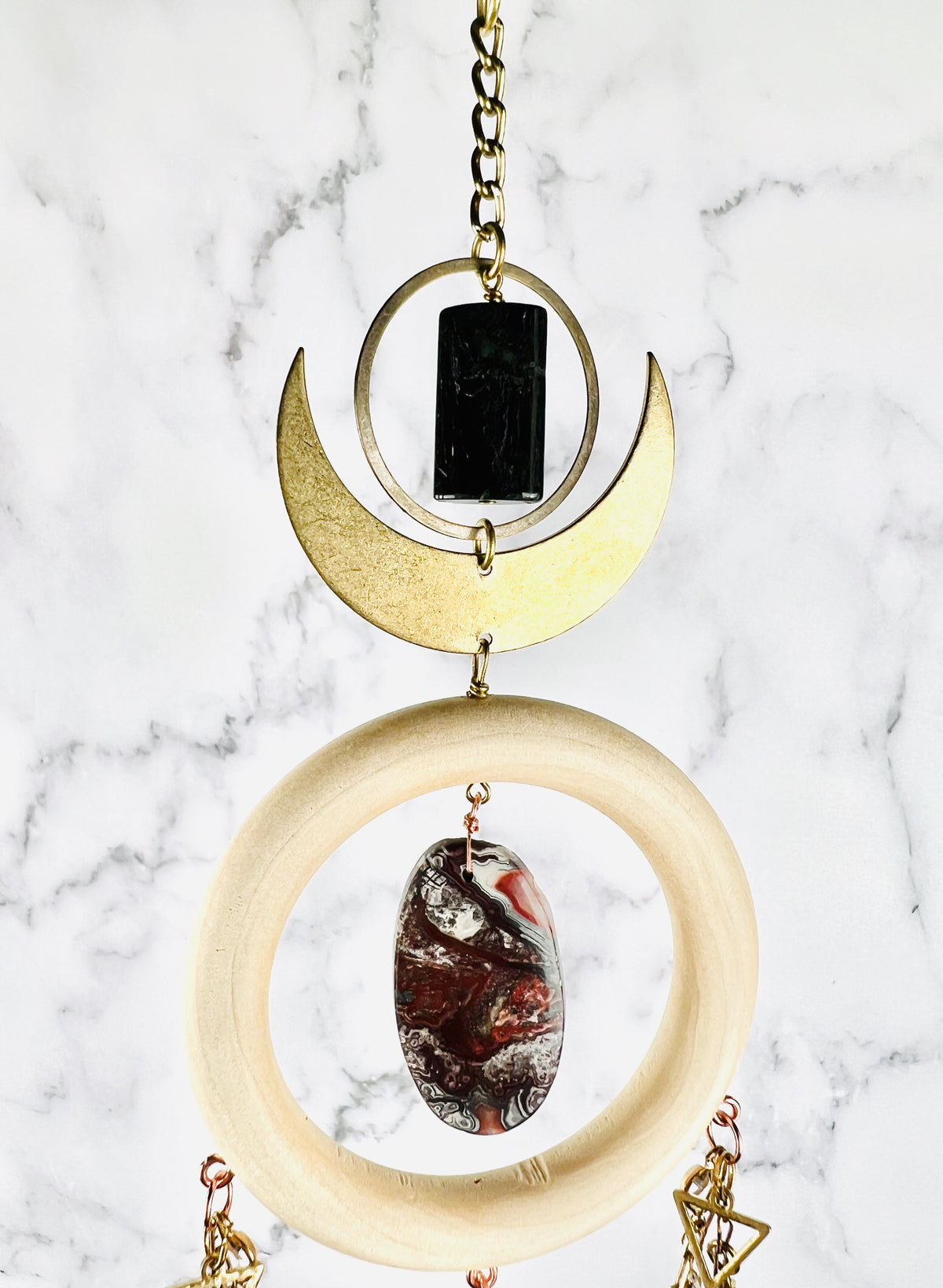 Depicted is a dimensional vertically hanging top portion of crystal wall art with gold chain, a bronze ring, and Crescent Moon, a wood ring with an center stone of red and gray crazy lace agate, with triangles and chain attached to the wood ring. All hangs in front of a gray and white marble wall.
