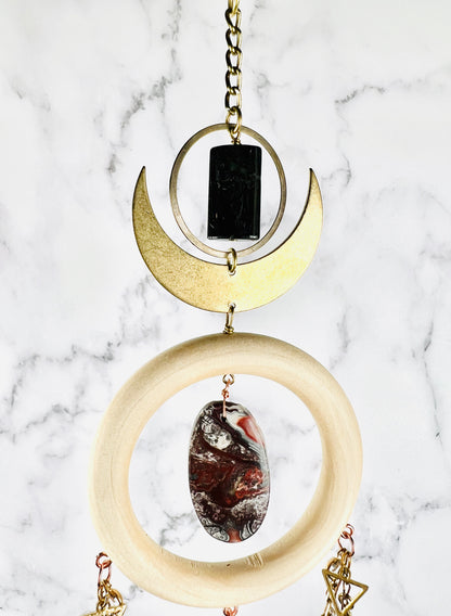 Depicted is a dimensional vertically hanging top portion of crystal wall art with gold chain, a bronze ring, and Crescent Moon, a wood ring with an center stone of red and gray crazy lace agate, with triangles and chain attached to the wood ring. All hangs in front of a gray and white marble wall.