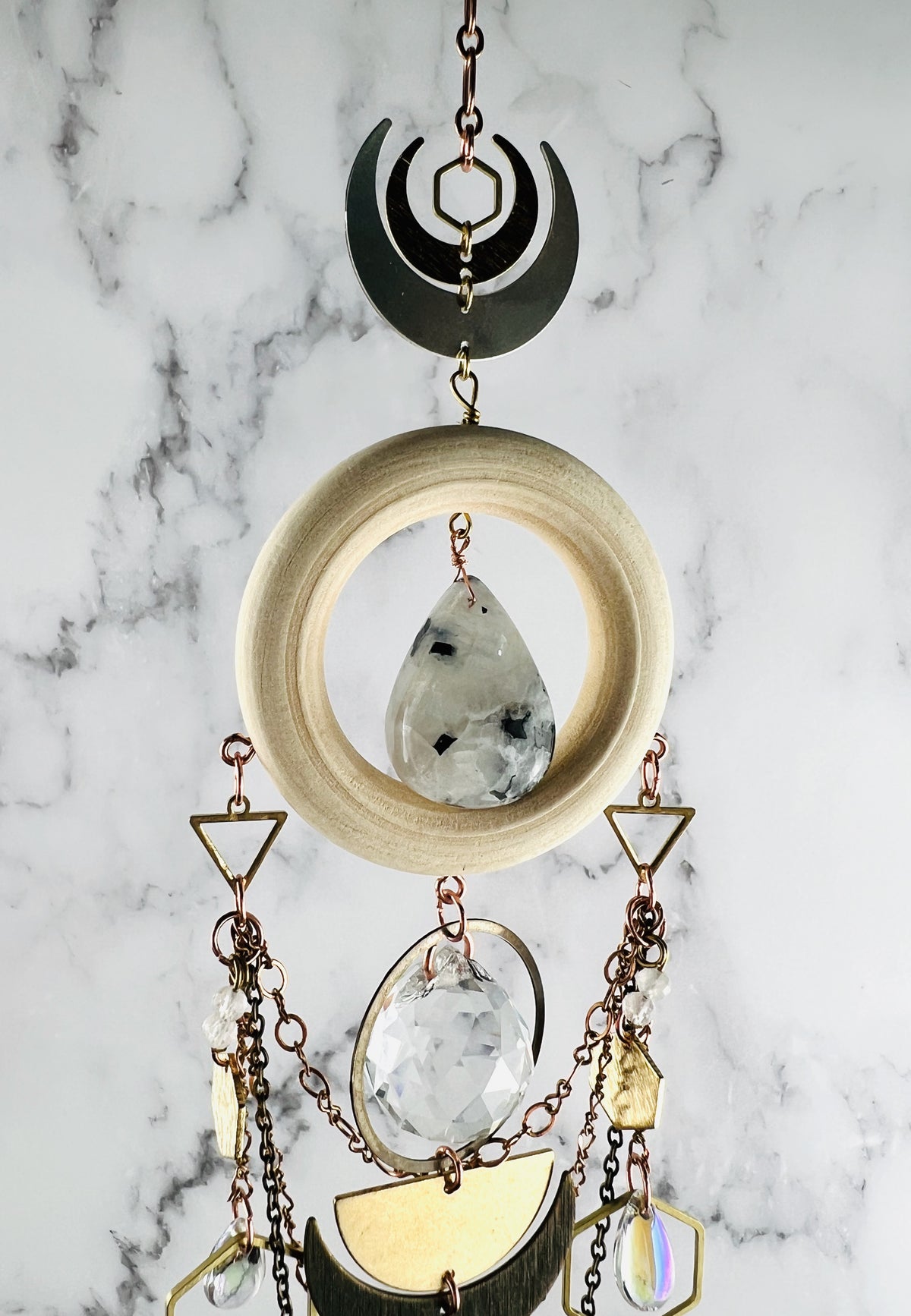 A beautiful vertically hanging crystal wall art piece includes: Copper chain hangs at the top and down from that are a hexagon and crescent moons attached to a light wood ring with a moonstone in the middle. Attached to the bottom of the wood are triangles, copper and bronze colored chain, rings, hexagons, and prisms, wire later today, wire wrapped beads, all hanging in front of a gray and white marble wall.