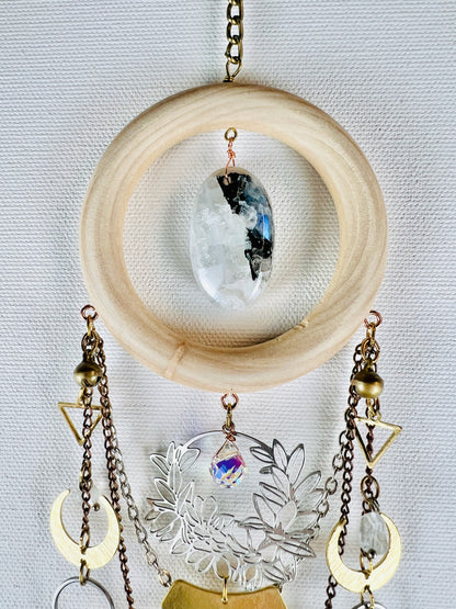 A bronze chain connected to a light colored wood ring with Center stone of black-and-white Moonstone, hangs on a light gray canvas with components, including: bronze and silver chain, brass bells, gold colored metal triangles, crescent moons, silver rings, and a silver flower wreath with blue and clear prism in the middle. Beautifully designed intentional home decor.