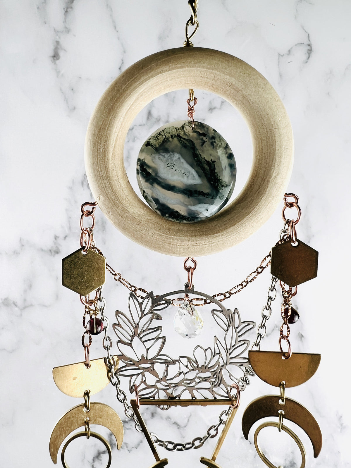 crystal wall art with bronze colored chain attached to a light wood ring with a circular green and white moss agate stone in the middle. Attached to the wood artfully are gold and silver colored chains metal, hexagons, beads, half moons, crescent moons, rings and a silver flower charm with a clear prism drop. This art hangs vertically in front of a gray and white marble background. 