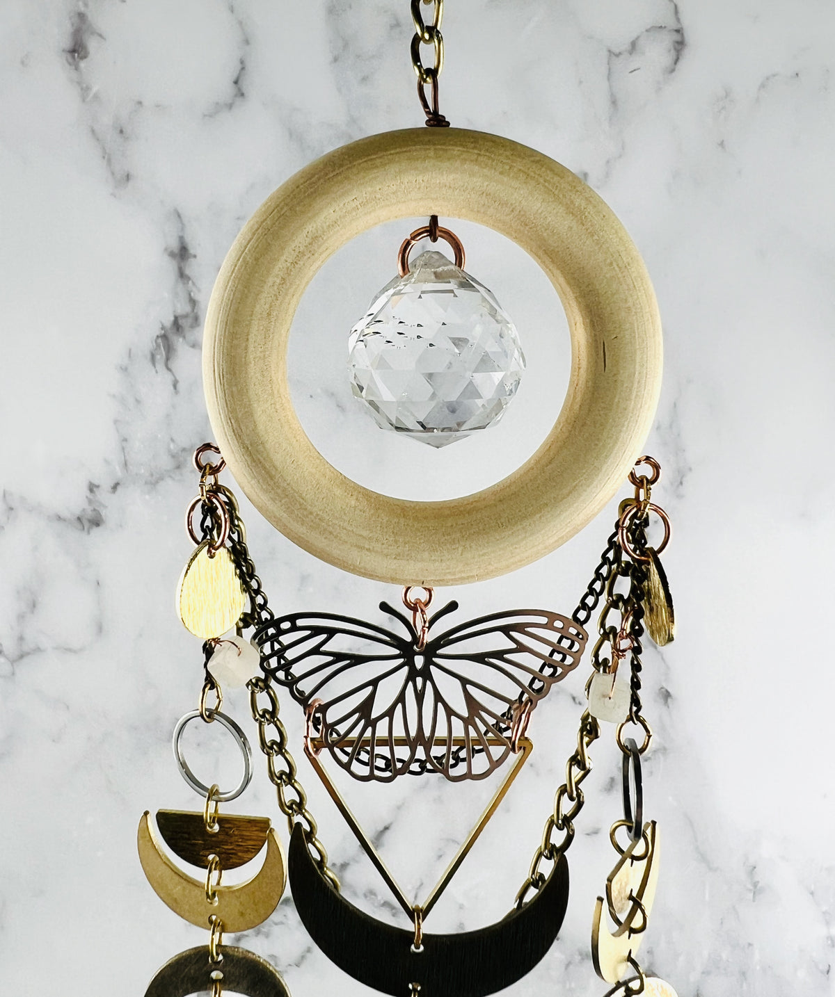top portion of vertically hanging crystal wall art with metal charms including a silver metal butterfly, a triangle, crescent moons, half circles, silver rings, gold colored chain and crystals on a marble background. Pink Aventurine and Topaz with large prism in the center of a light wood ring. A suncatcher and manifestation charm, created for intentional home decor and emotional balance