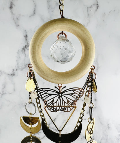 top portion of vertically hanging crystal wall art with metal charms including a silver metal butterfly, a triangle, crescent moons, half circles, silver rings, gold colored chain and crystals on a marble background. Pink Aventurine and Topaz with large prism in the center of a light wood ring. A suncatcher and manifestation charm, created for intentional home decor and emotional balance