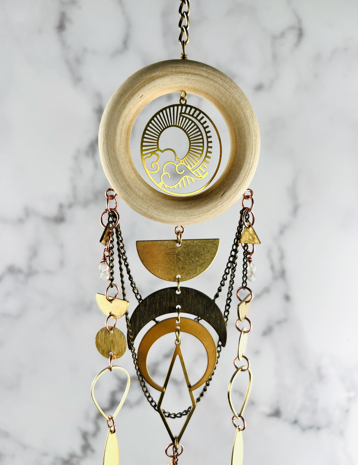 Top section of a piece of vertically hanging crystal wall art made of wood, citrine, moonstone, chain, a sun and clouds Charm, half circles, circles, triangles, crescent, moons, and diamond metal shapes; hanging in front of a marble wall.