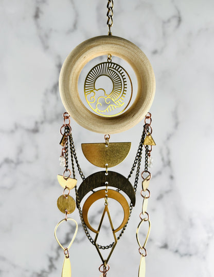 Top section of a piece of vertically hanging crystal wall art made of wood, citrine, moonstone, chain, a sun and clouds Charm, half circles, circles, triangles, crescent, moons, and diamond metal shapes; hanging in front of a marble wall.