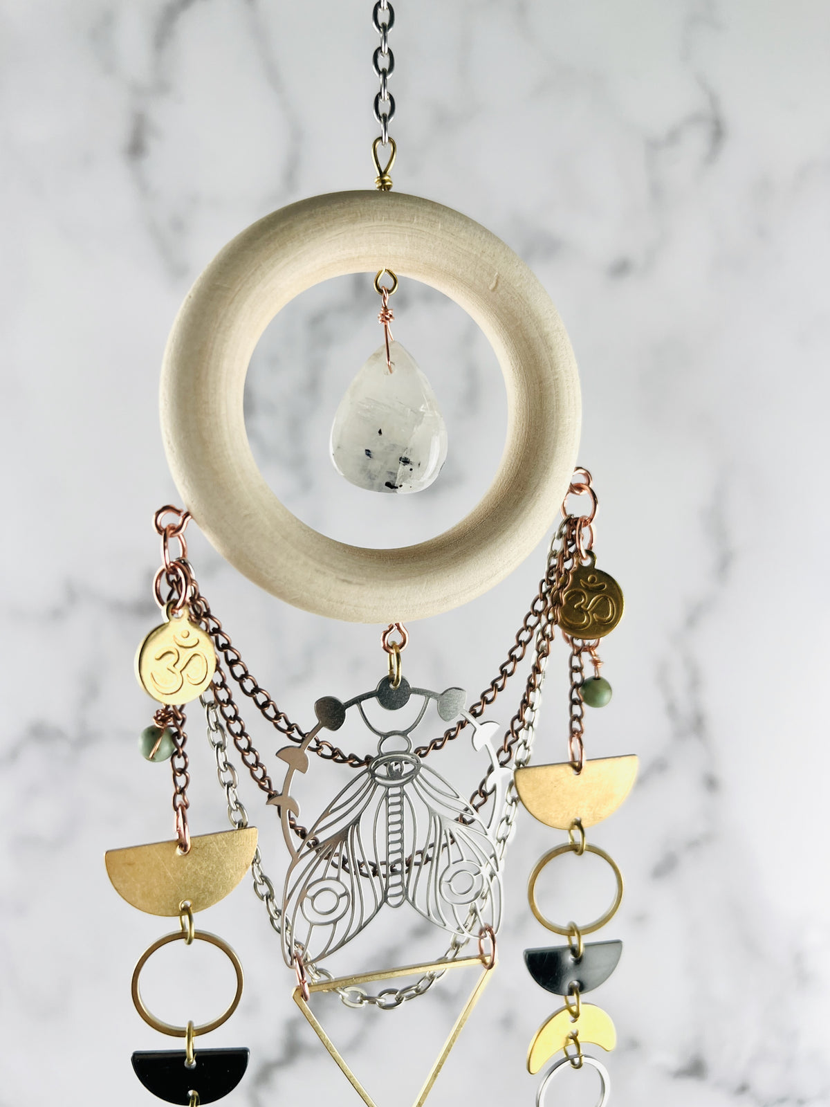 The top portion of a piece of crystal wall, art with silver chain at the top connected to a piece of cream, colored wood with a moonstone Drop shape in its center, and copper and silver colored chain with ohm symbols, light, blue wire, wrapped beads, half circles, gold rings, a triangle, crescent moon, and a silver moth with moon phase above it suspended in front of a gray and white marble wall.