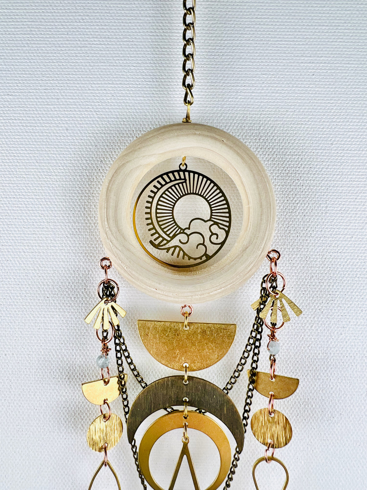 A light colored wood ring with sun, moon, and cloud, Charm hangs from a bronze colored chain in front of a light gray canvas wall. Connected and dangling from the wood ring, with copper components are gold colored metal fans, light blue aquamarine beads, half moons, crescent moons, circles, and swooping chains. All form and aesthetically pleasing piece of vertical, three-dimensional, art made of natural materials.