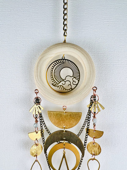 A light colored wood ring with sun, moon, and cloud, Charm hangs from a bronze colored chain in front of a light gray canvas wall. Connected and dangling from the wood ring, with copper components are gold colored metal fans, light blue aquamarine beads, half moons, crescent moons, circles, and swooping chains. All form and aesthetically pleasing piece of vertical, three-dimensional, art made of natural materials.