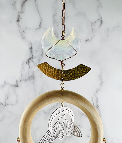 A piece of hanging crystal wall art, featuring a translucent iridescent crescent moon wrapped in copper wire, a hammered brass arch, and a light wood ring containing a silver-toned bee cut out with a moon phase graphic above it, all suspended by a copper chain hanging in front of a marble wall.