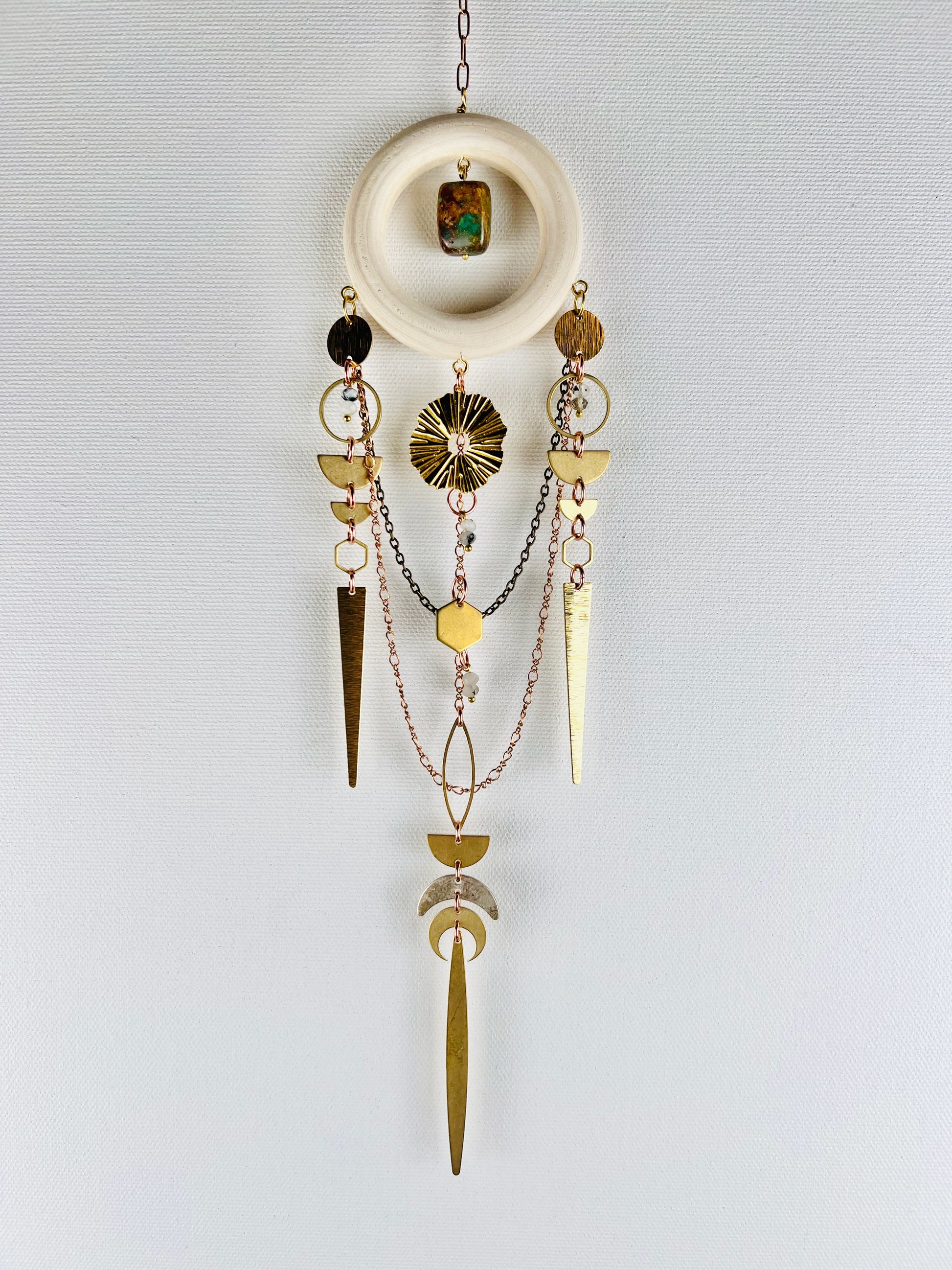 Decorative hanging crystal wall art with geometric shapes, such as sun burst, half circles, hexagon, crescent moons, circles, spikes, and eye shapes. also has turquoise, moonstone, chain and a wood ring, on a light gray background