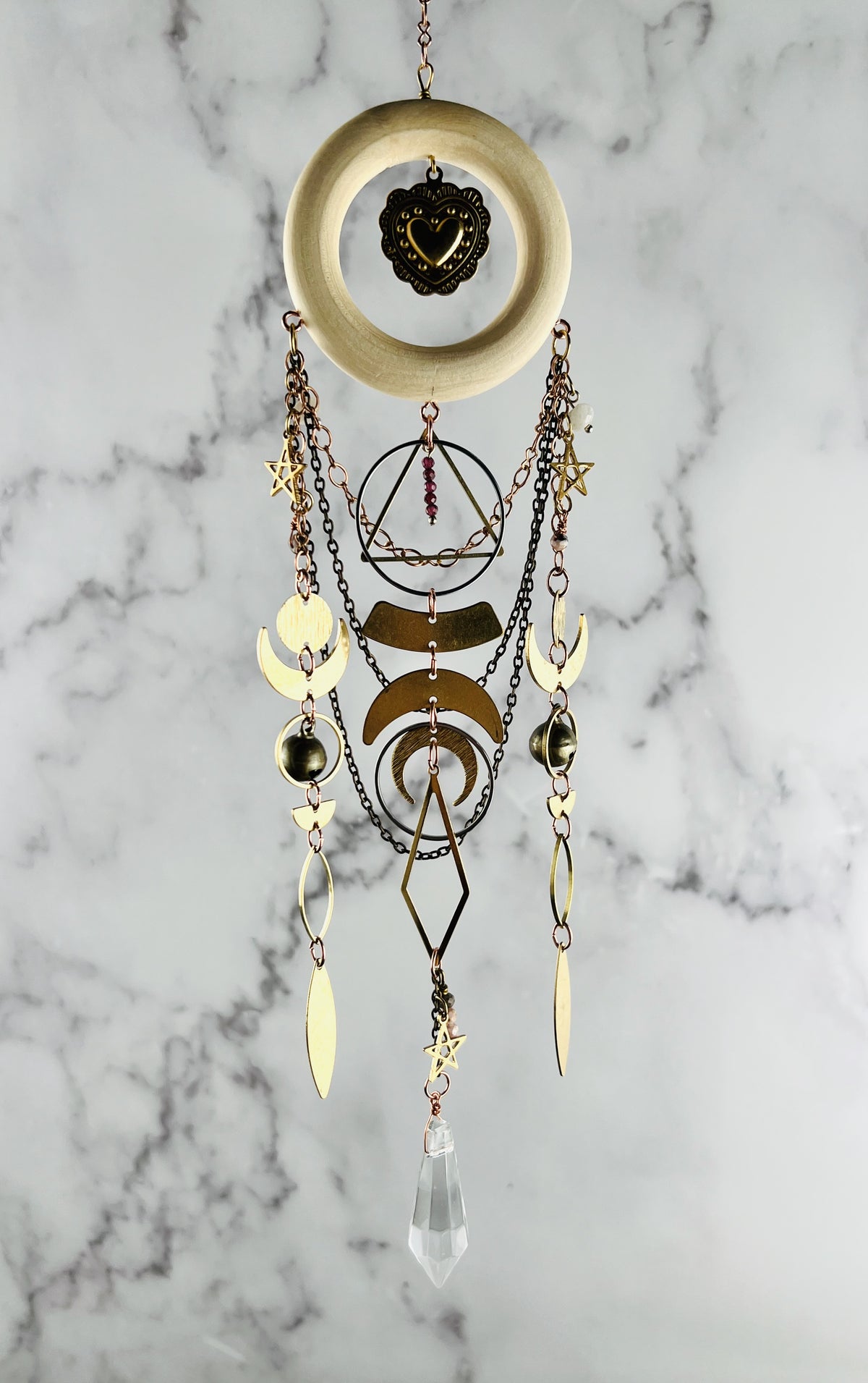 Light wood ring hanging from a chain at the top with chains also hanging from the bottom of it, as well as various gold, geometric shapes, including: circles, crescent, moons, rings, diamonds, stars, triangles with three chains hanging from it as well. There are also red garnets and pink adventuring beads hanging from this. piece of art and it is hanging in front of a marble wall.