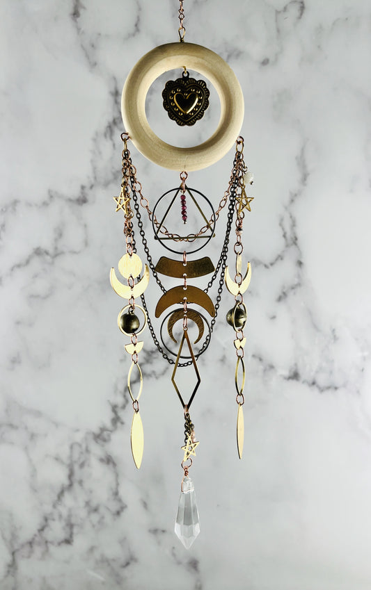 Light wood ring hanging from a chain at the top with chains also hanging from the bottom of it, as well as various gold, geometric shapes, including: circles, crescent, moons, rings, diamonds, stars, triangles with three chains hanging from it as well. There are also red garnets and pink adventuring beads hanging from this. piece of art and it is hanging in front of a marble wall.